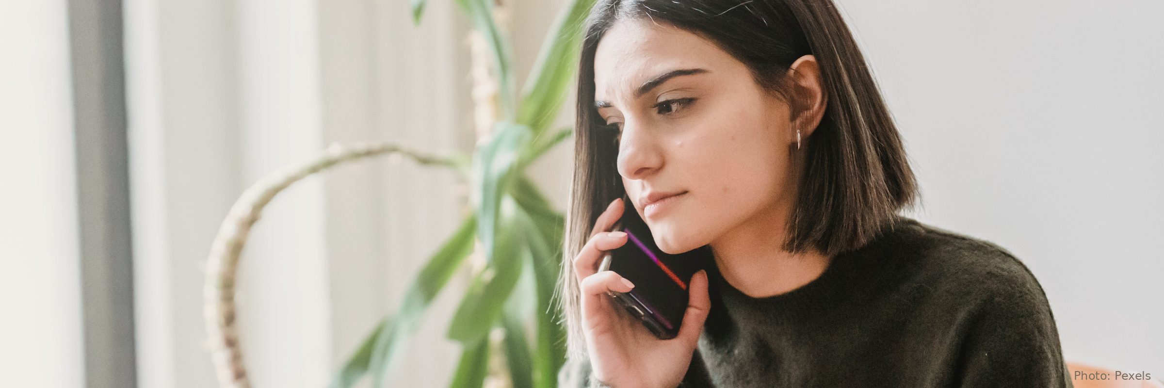 A woman sits at home by a bright window, making a phone call.