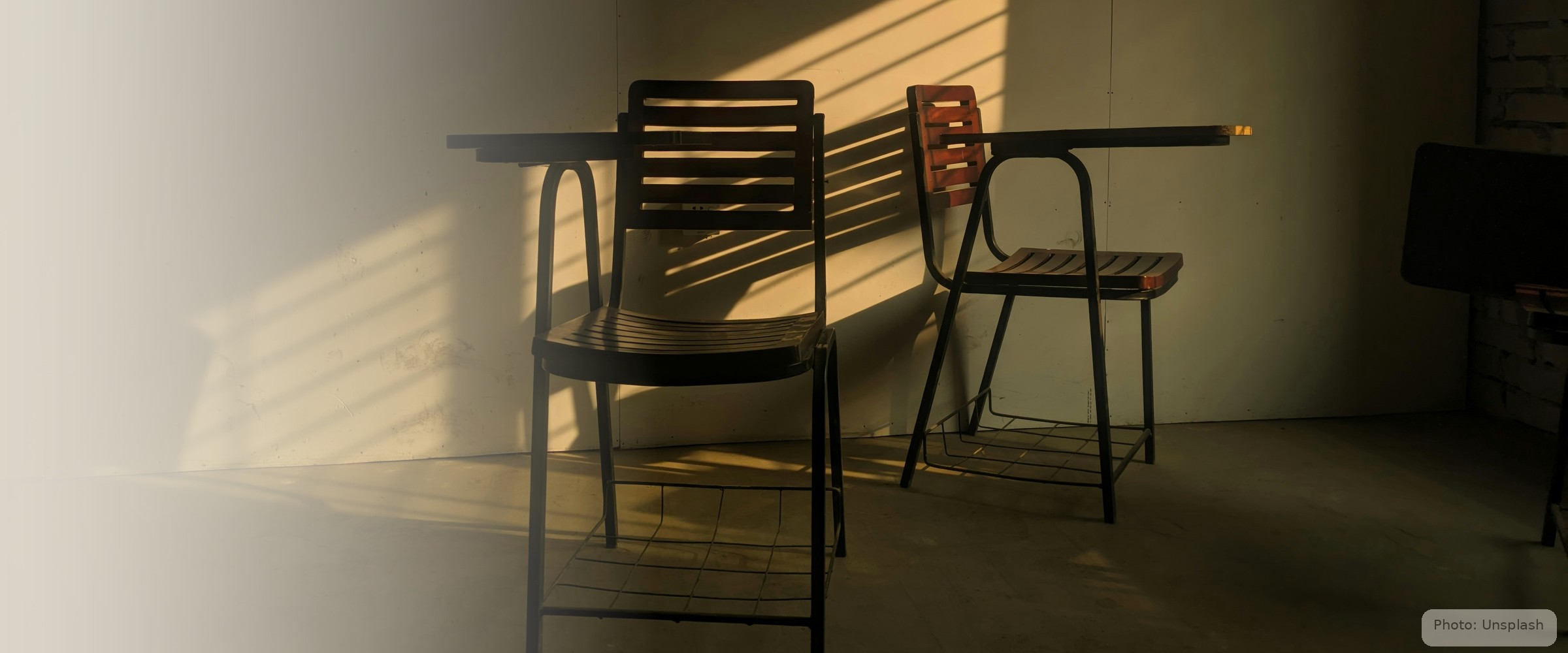 Two empty chairs catch a shaft of late light against a bare wall — the furniture of absence.