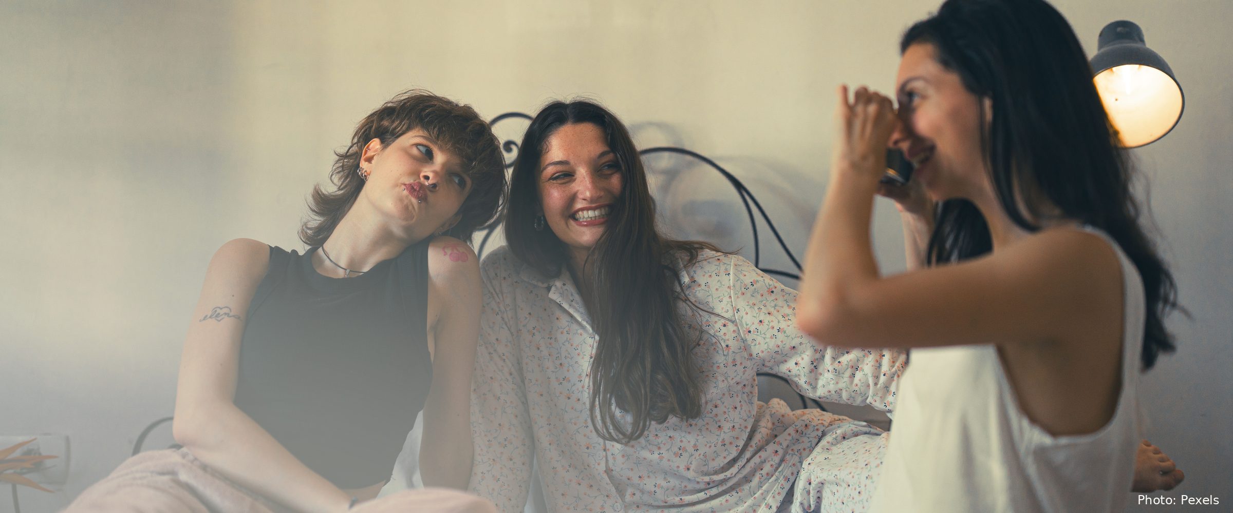 Three women sit close together on a bed, laughing in warm lamplight.